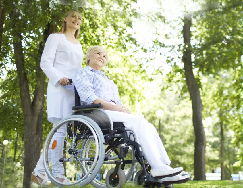 A Woman Helping An Elderly Lady Patient Sitting On A Wheelchair In A Park